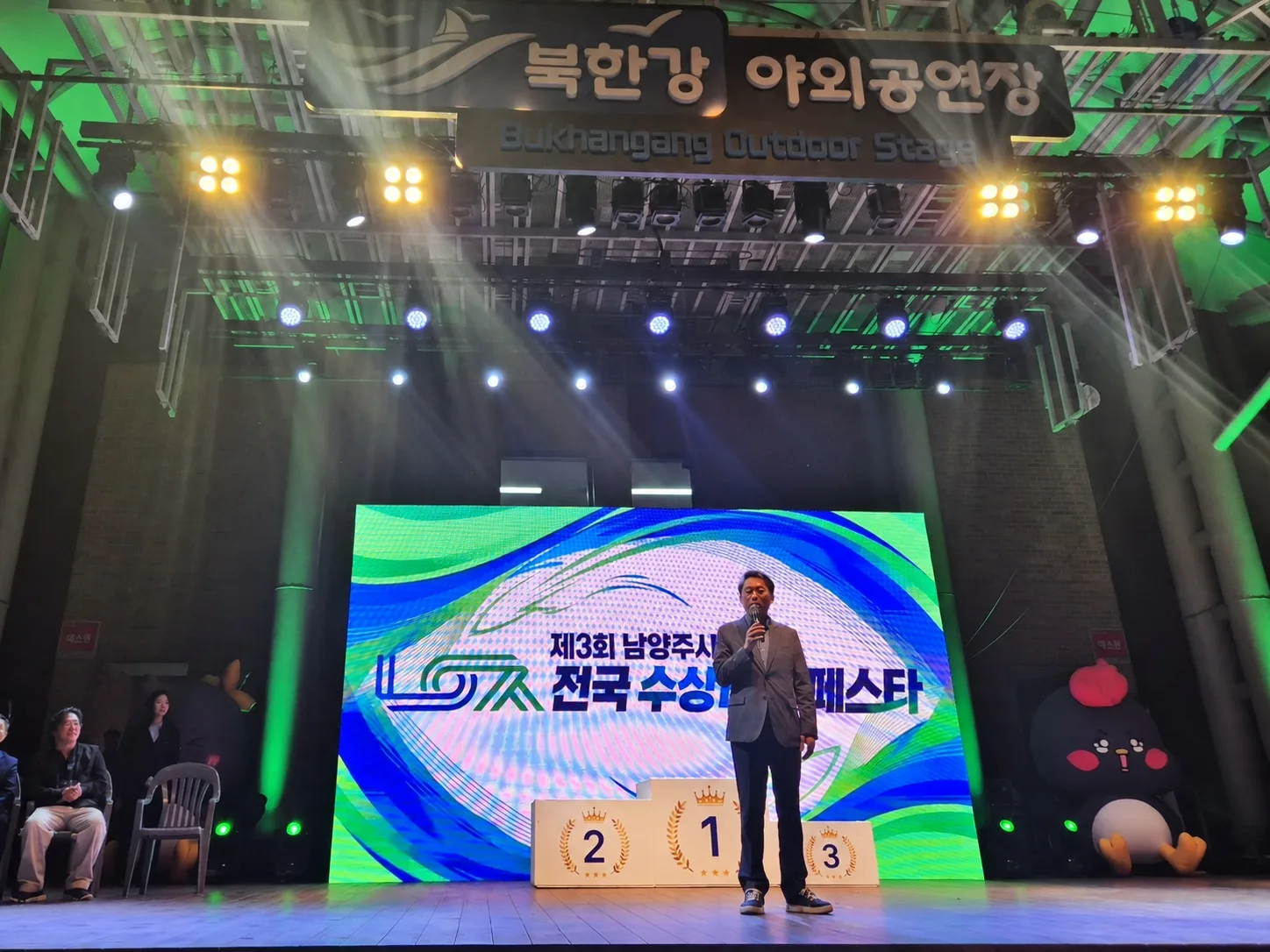 무대 위에 시상대가 놓여있고 스크린에 행사 이름이 보이는 가운데 한 남성이 무대 중앙에서 발표하거나 연설하는 장면