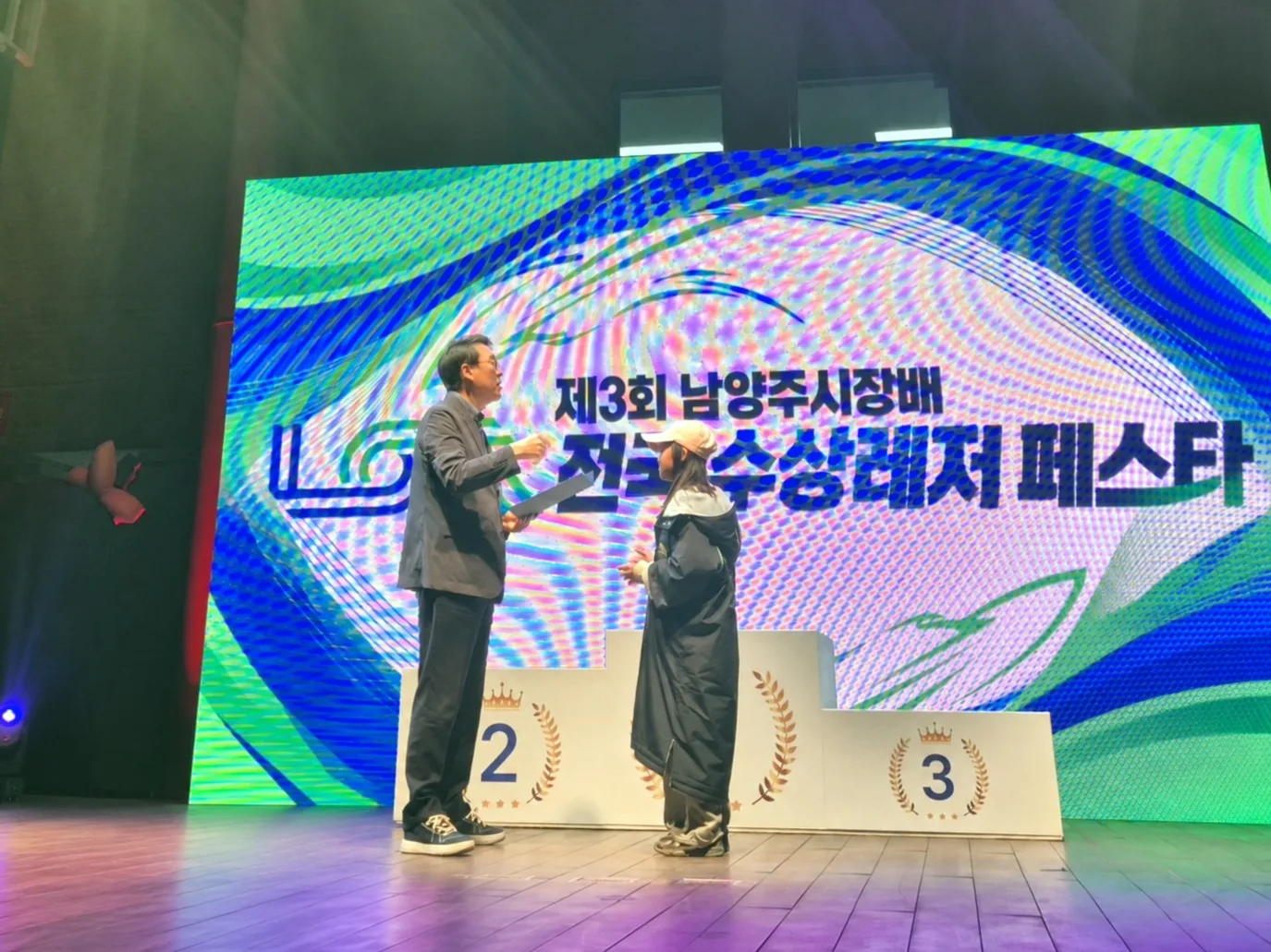 무대 앞에서 두 사람이 마이크를 들고 대화하거나 공연 준비를 하는 모습, 뒤쪽에 큰 스크린에 행사명이 보임