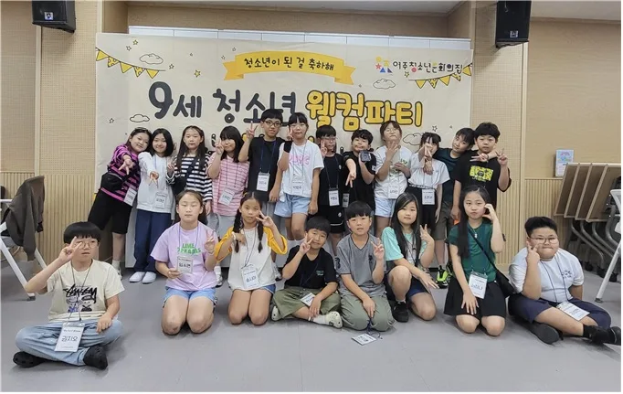 9세 어린이들이 '9세 청소년 웰컴파티' 행사 현장 앞 배너를 배경으로 다 함께 모여 앉거나 서서 손으로 평화 표시를 하며 단체 사진을 찍고 있는 모습