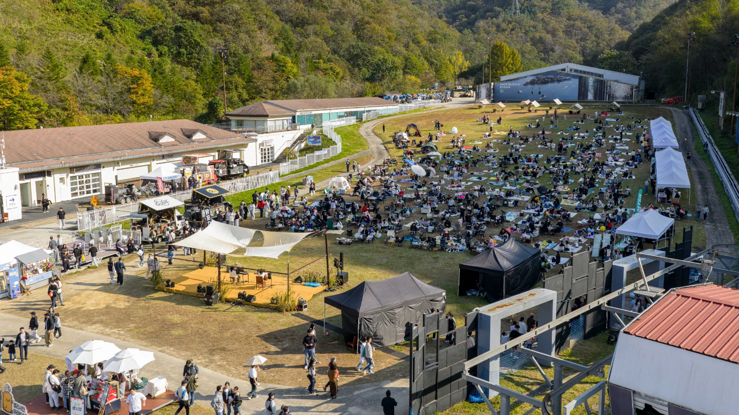 가을 산자락 아래 넓은 잔디밭에 여러 개의 텐트와 돗자리들이 펼쳐져 있고 사람들이 산책하고 있는 모습