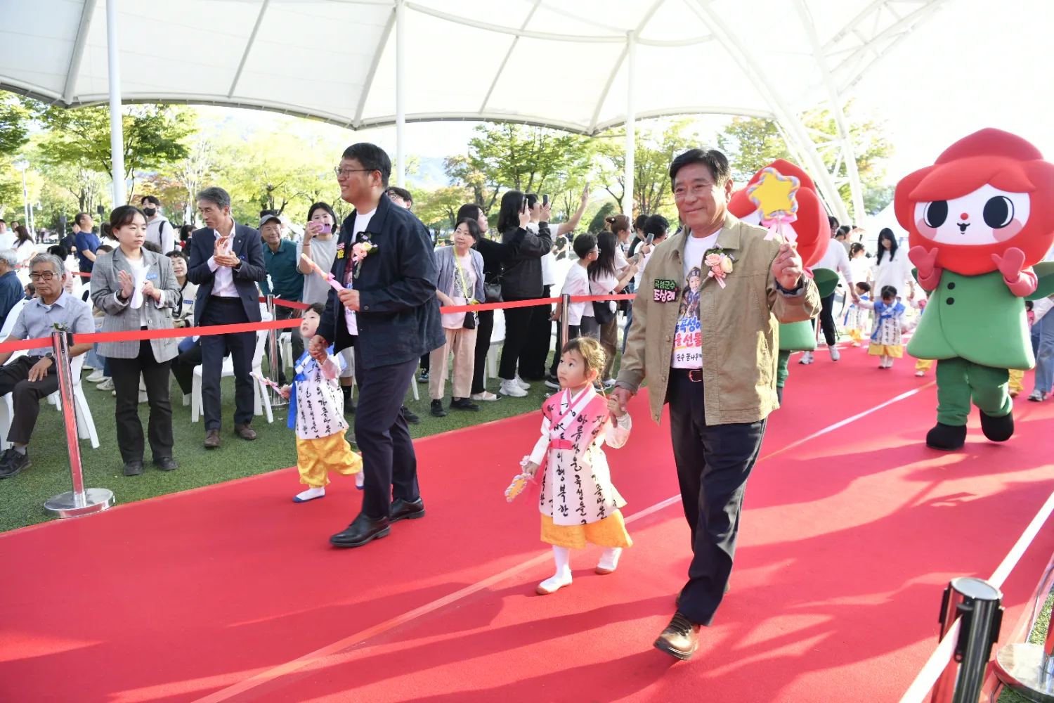 붉은 카펫 위를 따라 어린이와 성인들이 손을 잡고 걷고 있으며, 캐릭터 탈을 쓴 인물이 옆에서 함께 걷고 있는 모습.