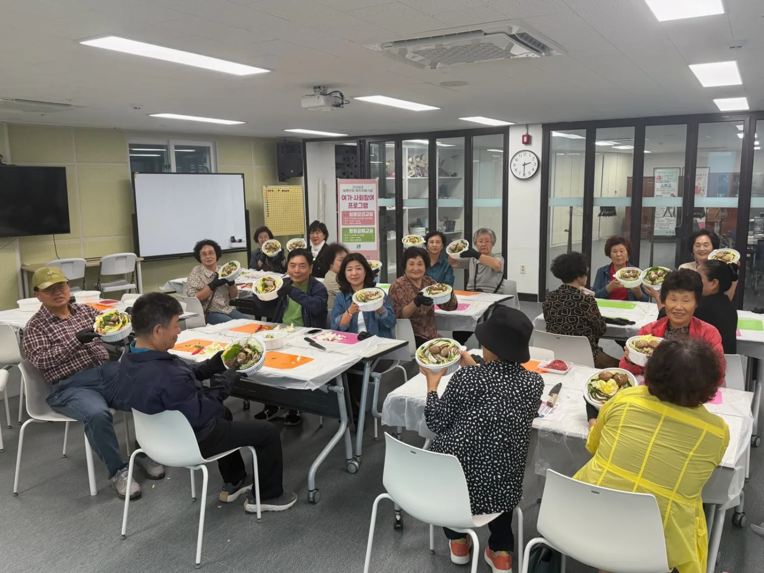 실내 강의실에서 여러 명의 어르신들이 각자 접시에 담긴 밀푀유나베 요리를 앞에 두고 앉아 카메라를 향해 미소 지으며 보여주고 있는 모습