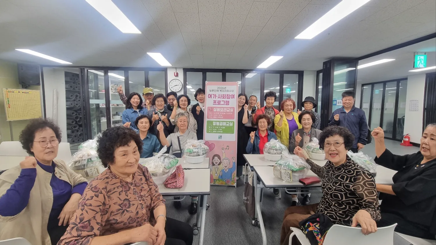 실내 강의실에서 20명가량의 어르신과 진행자가 프로그램 안내 배너 앞에서 손가락 하트 포즈를 취하며 밝게 웃고 있는 단체사진