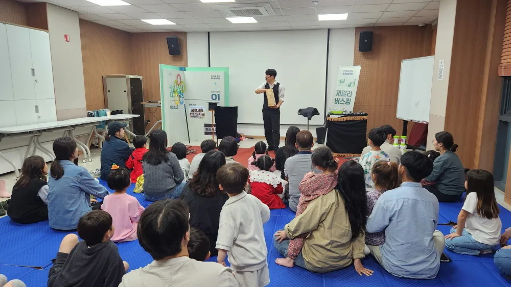 마술사가 무대에서 공연을 펼치고 앞에 모여 앉은 아이들과 가족들이 집중해서 관람하는 장면