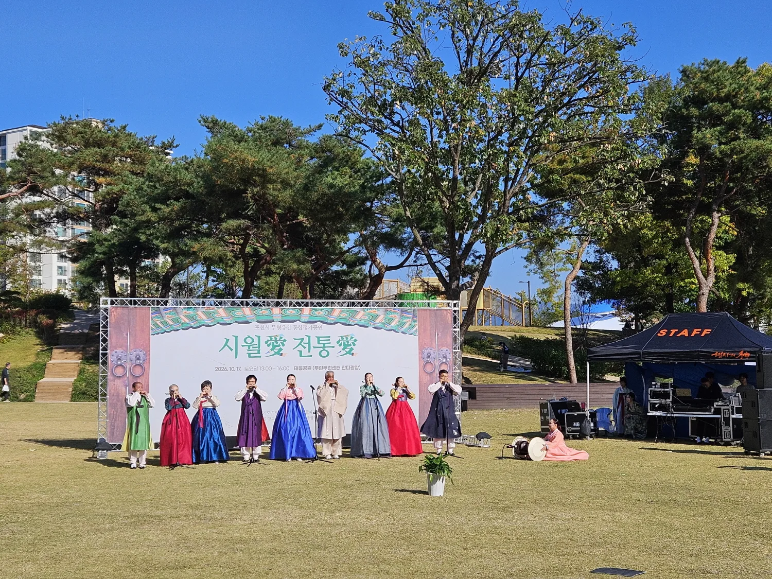잔디광장 무대 앞에서 전통 한복을 입은 여러 명의 공연자가 나란히 서서 풀피리를 부는 모습과 공연 준비 장비가 보이는 장면