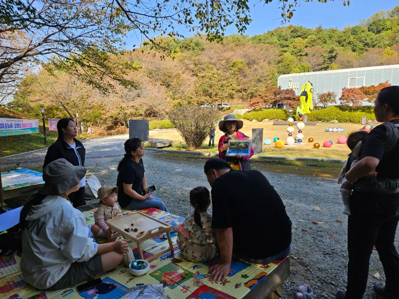 야외에서 모자와 가벼운 옷차림의 성인과 아이들이 둘러앉아 숲놀이 활동을 하며, 한 강사가 사진 자료를 보여주며 이야기를 하는 장면.