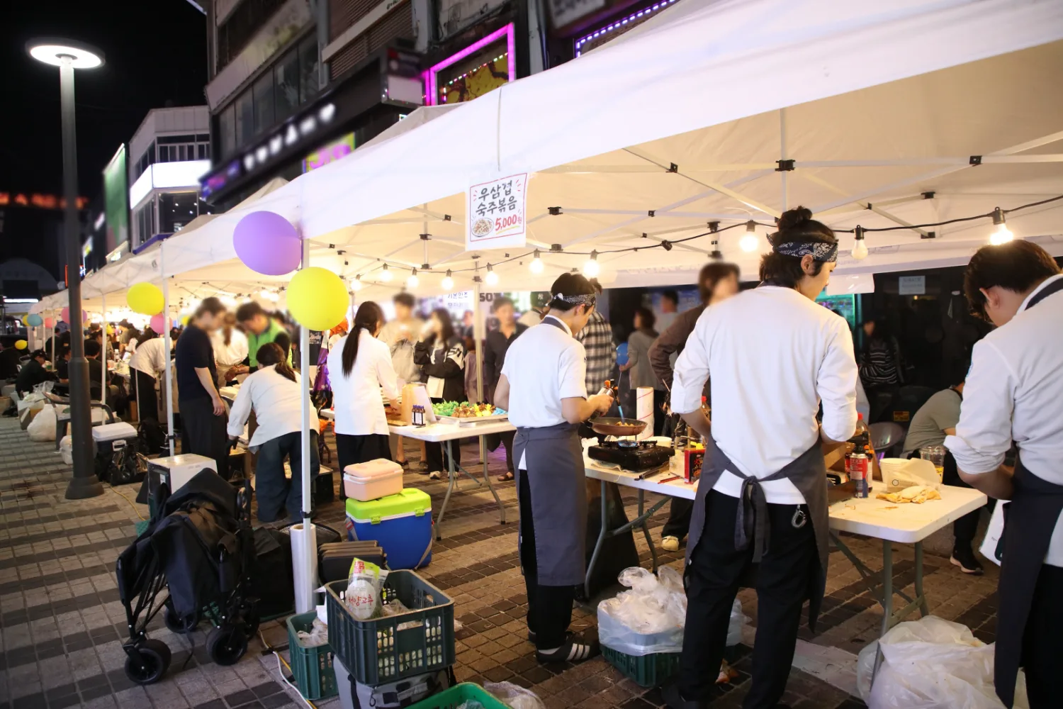 야외 천막 아래에서 조리복을 입은 직원들이 음식을 준비하고 방문객들이 줄 서서 기다리는 장면