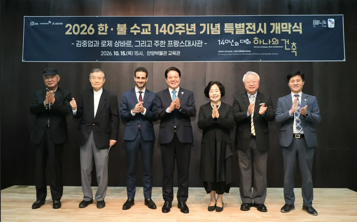 무대 앞에서 양복 차림의 일곱 인물이 나란히 서서 박수치는 모습의 단체 사진.