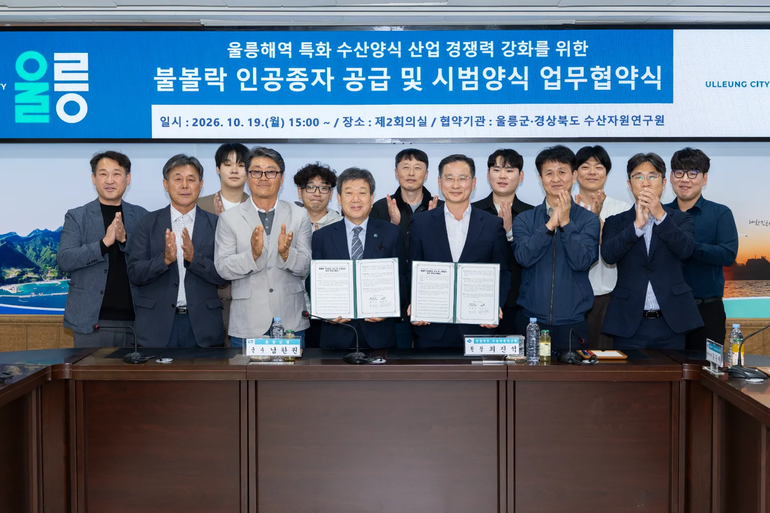 업무협약식에서 12명의 관계자가 나란히 서서 카메라를 향해 박수를 치며, 가운데 두 대표자가 업무협약서 두 권을 들어 보이고 있다.