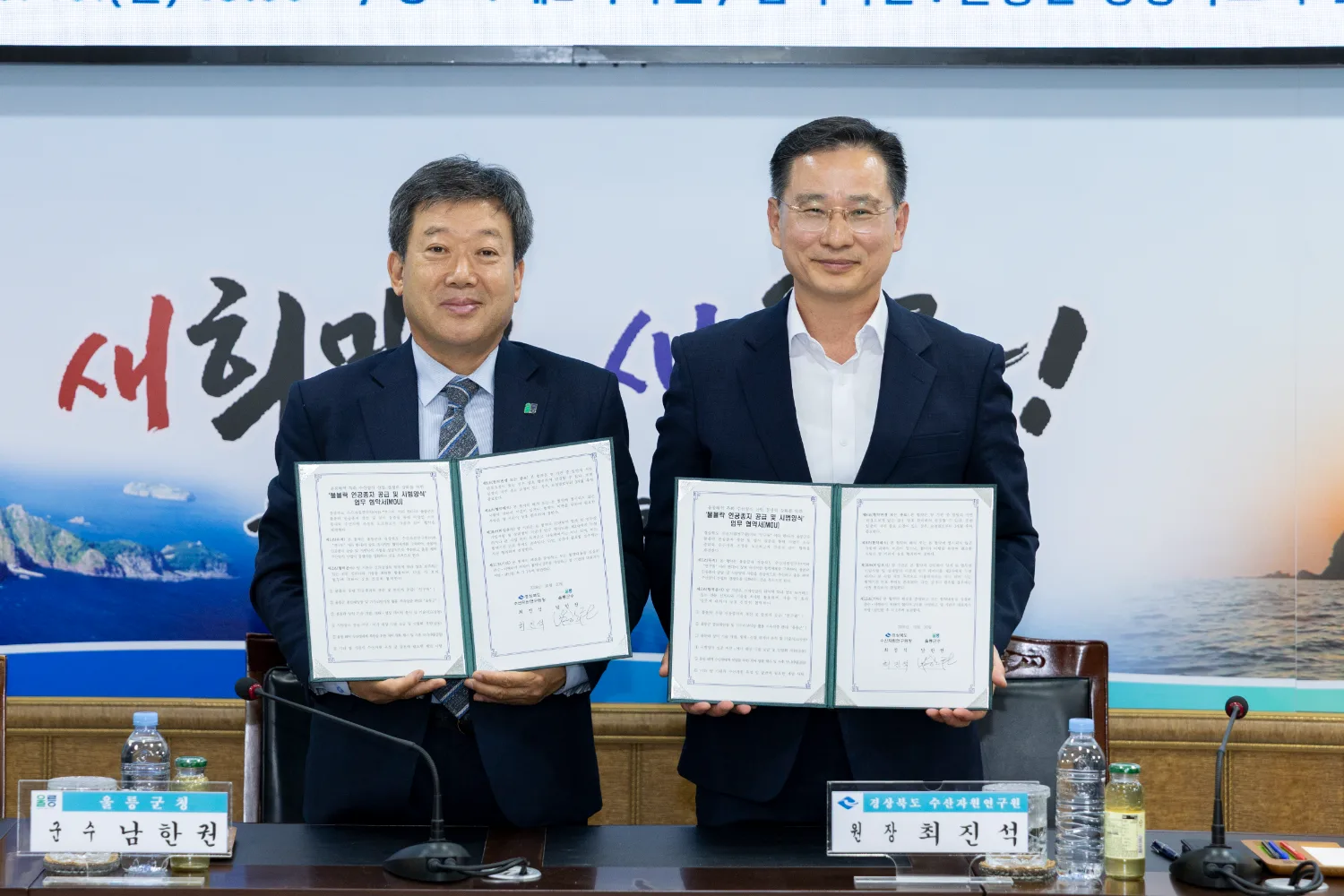 업무협약식에서 두 명의 남성이 협약서 두 권을 들어 보이며 웃는 모습.