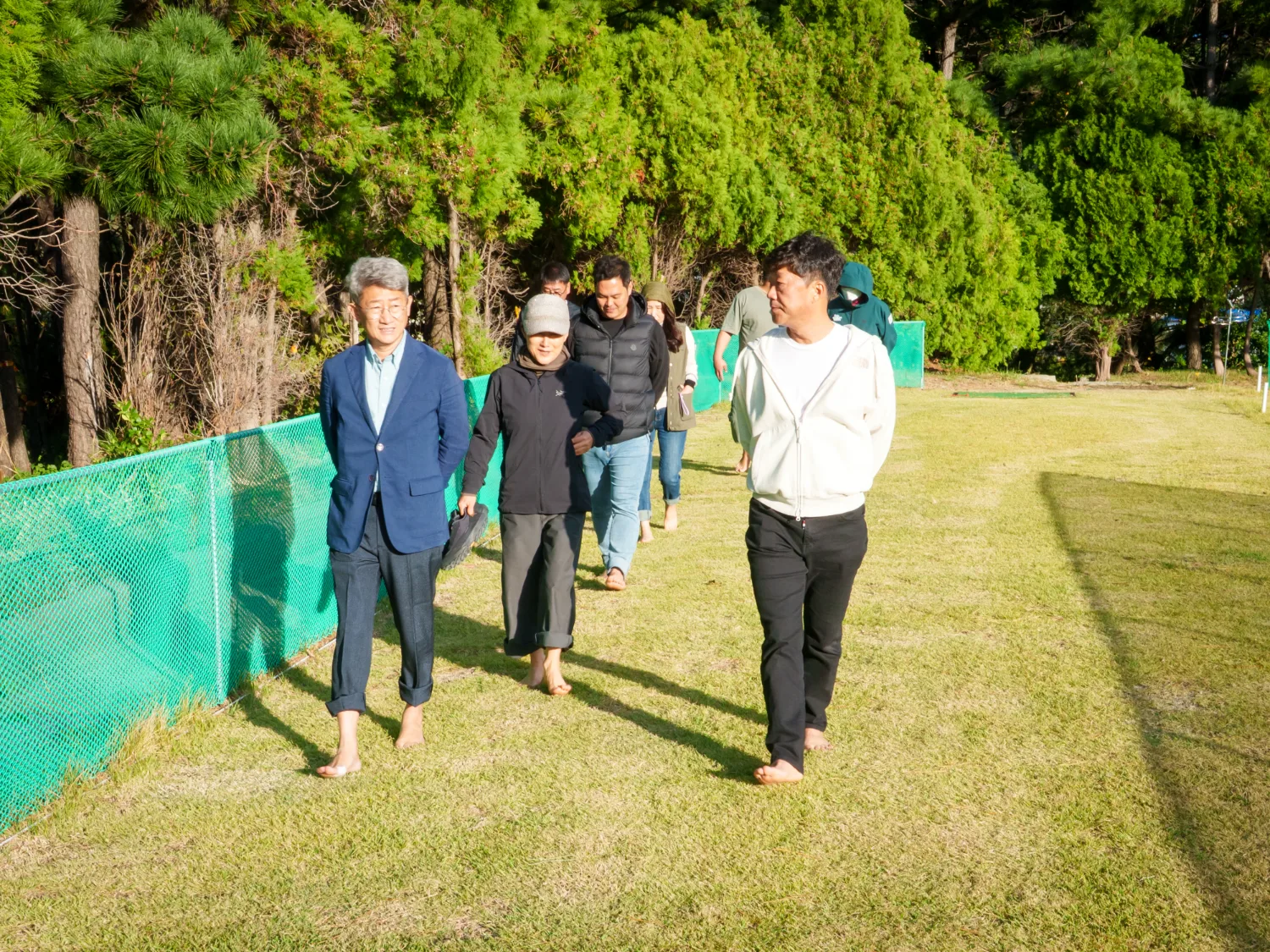넓은 잔디밭에서 여러 참가자들이 맨발로 걷는 모습으로, 뒷배경에 키 큰 나무들이 드리워져 있다.