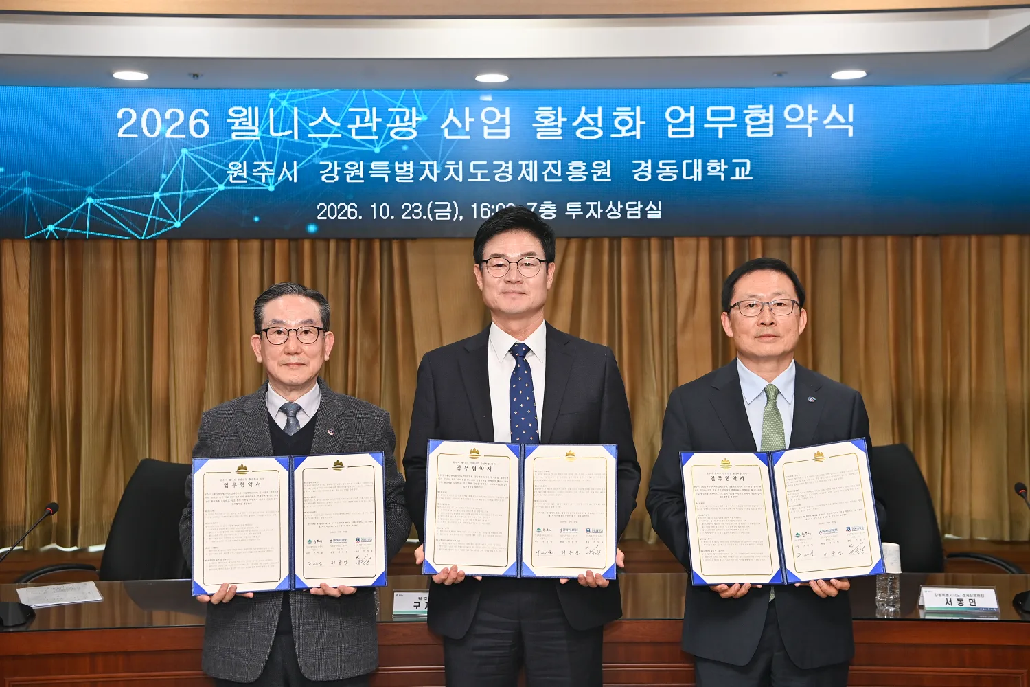 책상 앞에서 세 명이 업무협약서를 펼쳐 들고 있는 모습