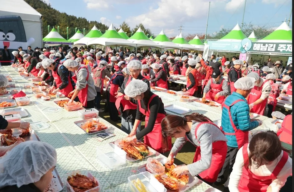 야외 행사장에서 일정한 제복을 입고 위생망을 쓴 다수의 사람들이 김치 만들기 체험을 하는 장면