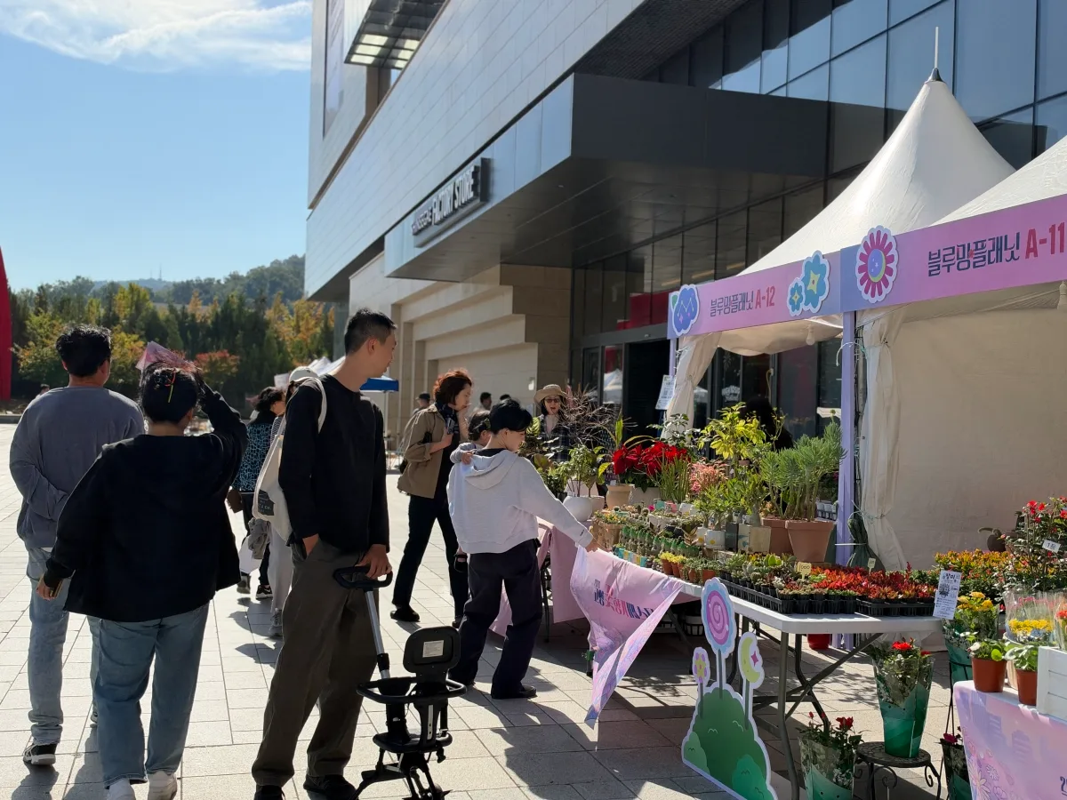 테이블에 진열된 다양한 화분과 식물을 보고 있는 가족 단위 방문객 모습, 야외 장터 분위기