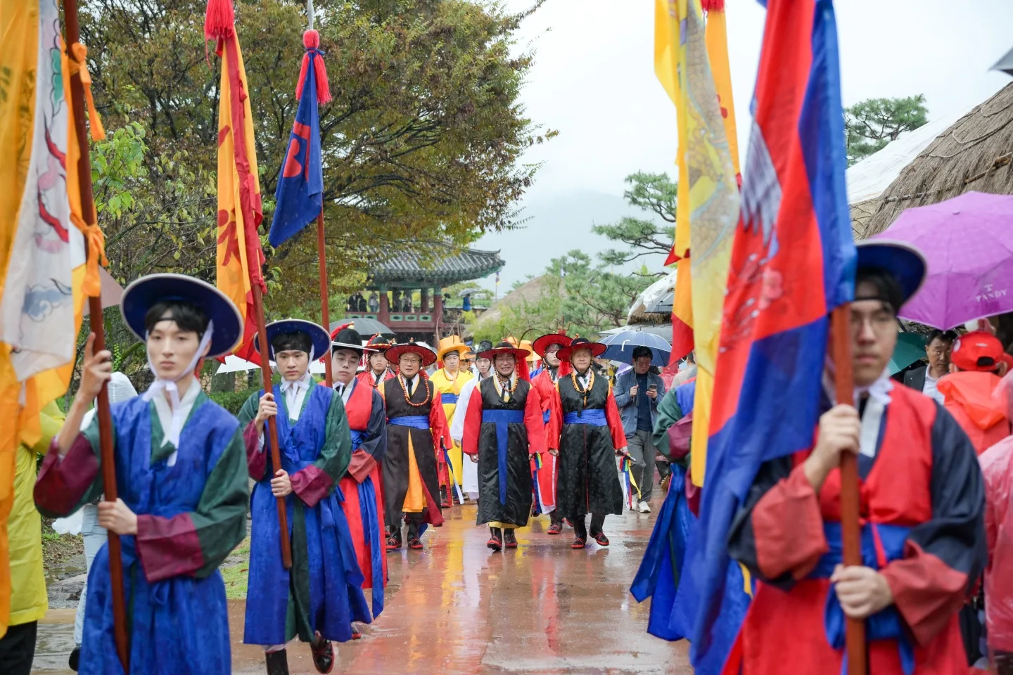 비 오는 날 낙안읍성 돌길에서 조선시대 전통 의상을 입은 사람들이 깃발을 들고 행렬을 하는 모습. 참가자들이 우산을 쓰거나 비를 맞으며 이동 중이다.