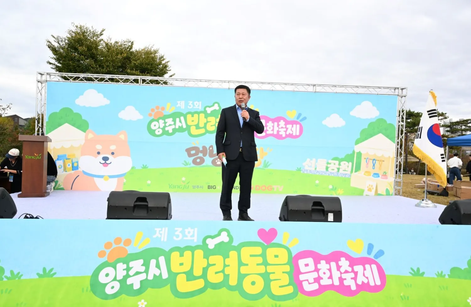 야외 무대 위에서 정장을 입은 인물이 마이크를 들고 연설하는 모습, 무대 배경에는 축제 이름이 크게 표시되어 있다.