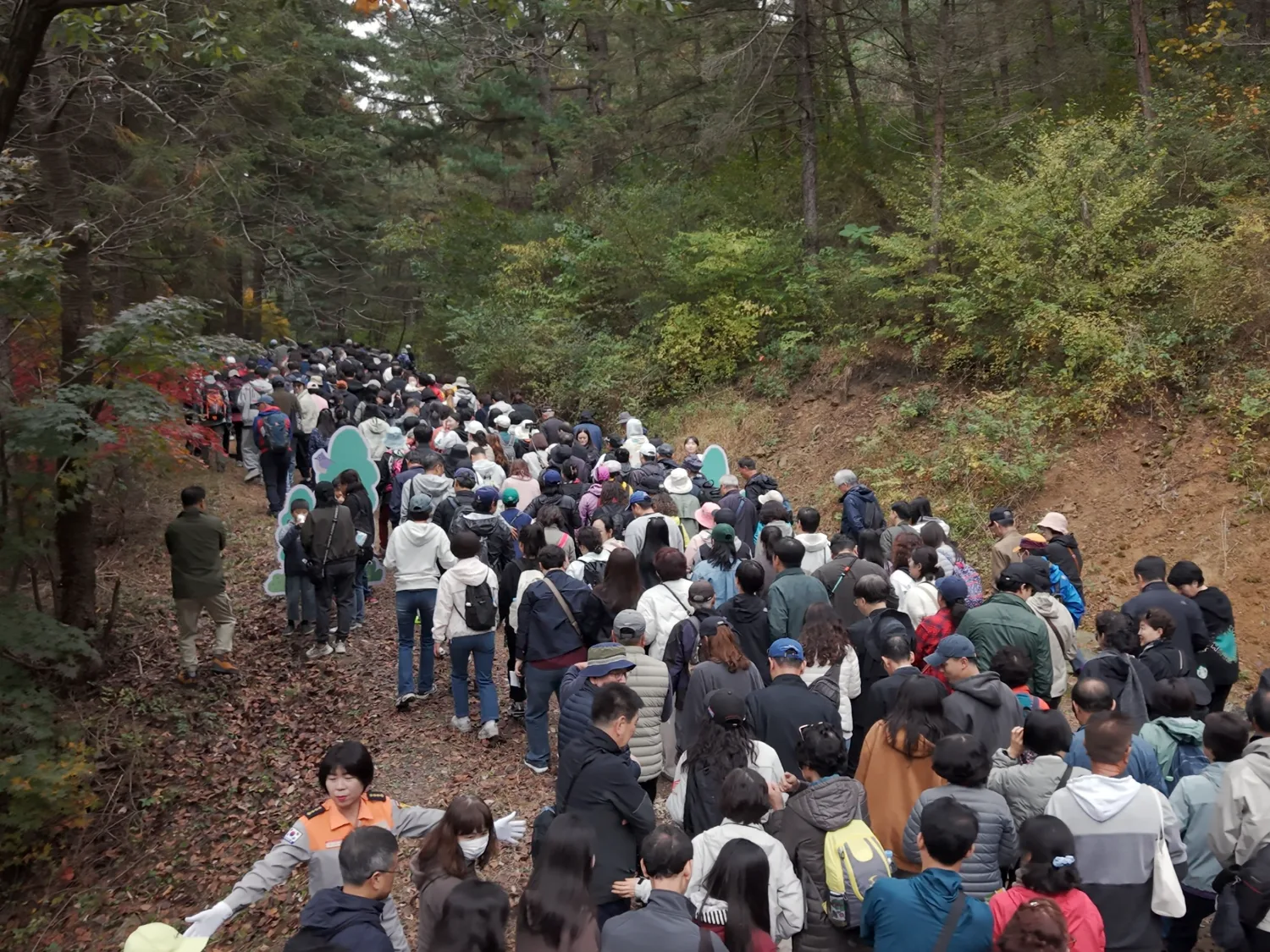 가을 단풍과 나무들이 울창한 숲길을 많은 사람이 한 줄로 걸어가고 있는 전경.