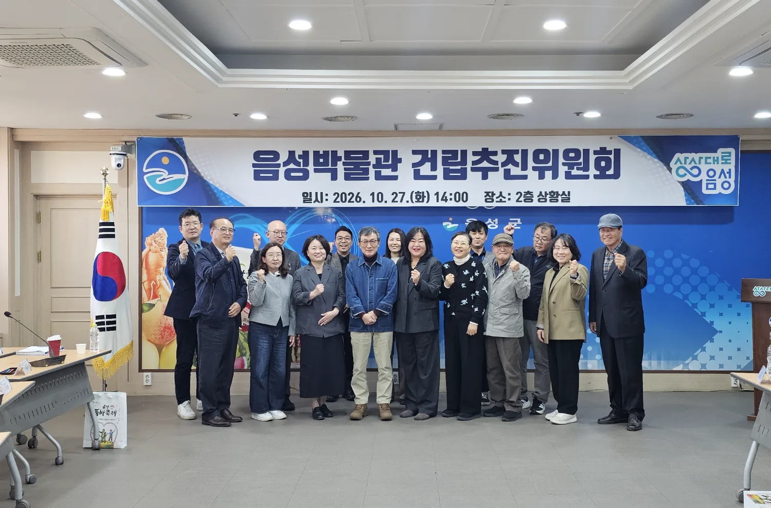 회의실에서 음성박물관 건립추진위원회 현수막 앞에 나란히 서서 손가락 하트를 만드는 15명의 중년 및 노년 남녀 위원들 모습