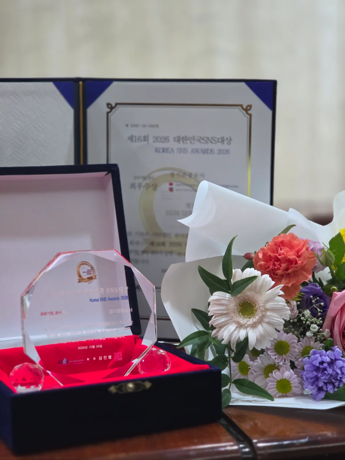 상패 두 개와 상패가 담긴 검은색 케이스, 그리고 색색의 꽃으로 구성된 꽃다발이 테이블 위에 놓여 있는 모습