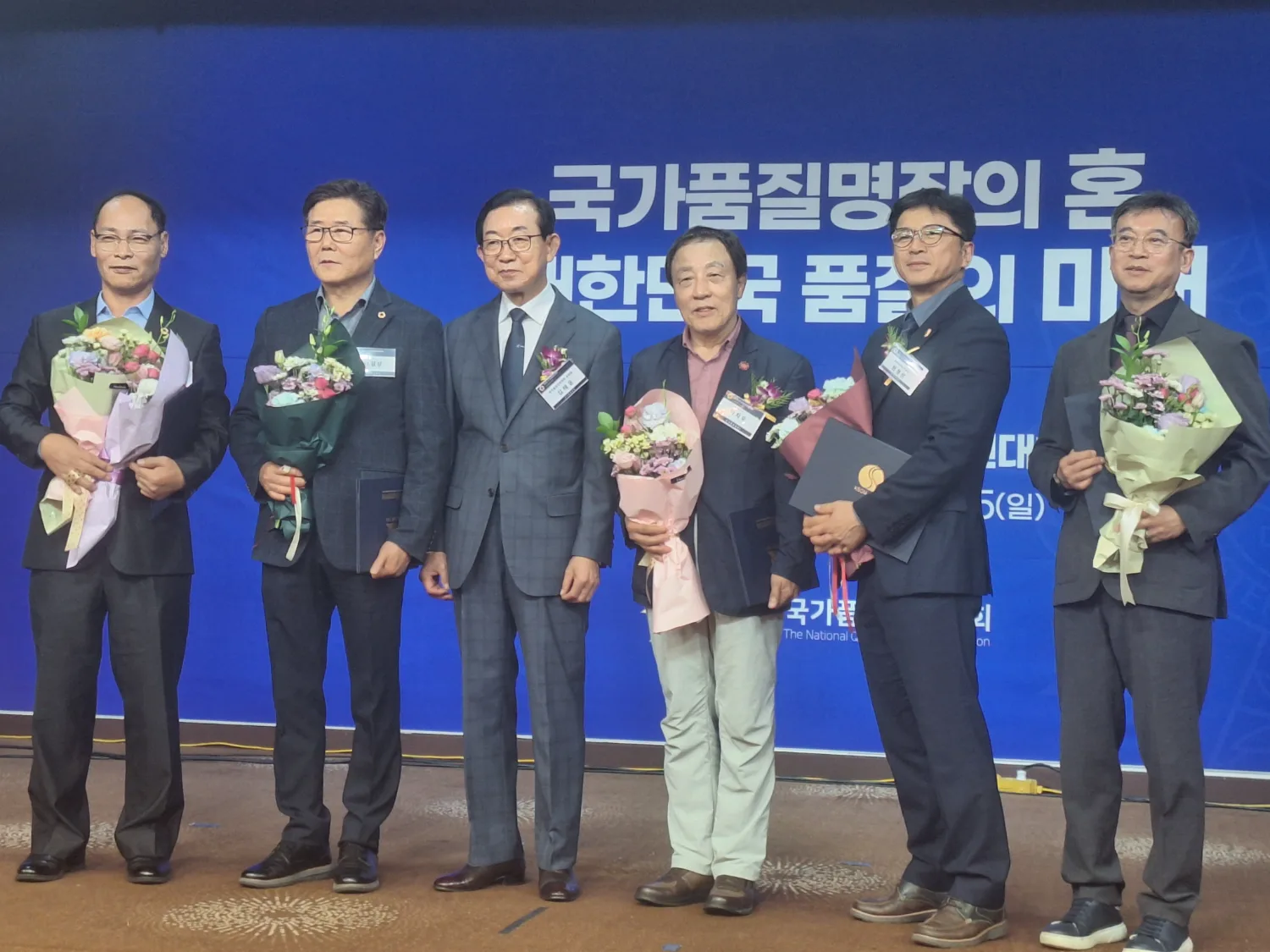 한국품질명장 수상자 여섯 명이 행사 배경 앞에서 꽃다발과 상장을 들고 서 있는 모습