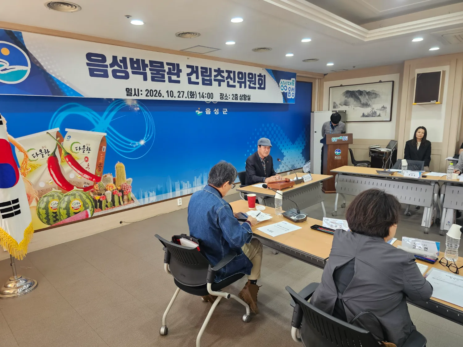 회의실에서 책상에 앉아 자료를 보는 위원들과 발표하는 여성, 배경에 음성군 로고와 박물관 건립 현수막이 있는 회의 장면