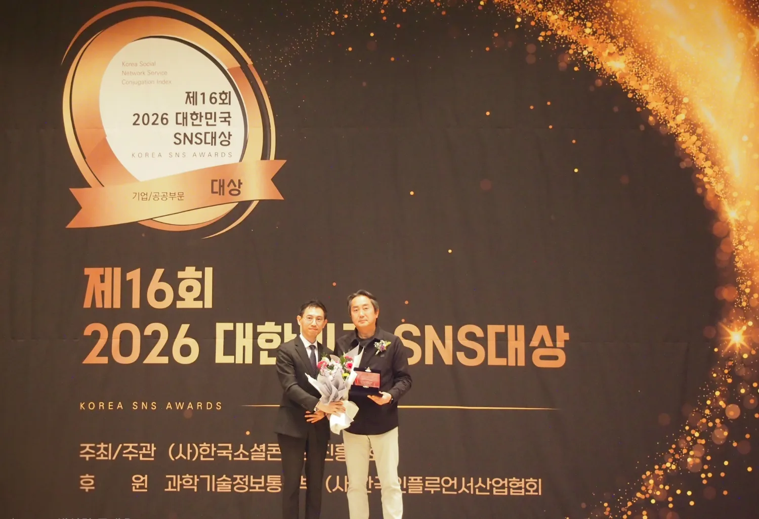 ‘2026 대한민국 SNS 대상’ 시상식 무대에서 두 명의 남성이 트로피와 꽃다발을 들고 기념 촬영을 하는 모습