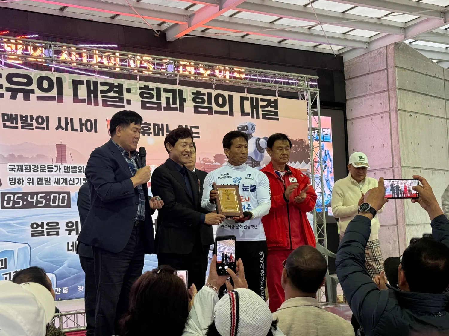 실내 공연장 무대에서 조승환 씨와 4명의 관계자가 상패를 들고 카메라를 향해 서 있고, 관중들이 사진을 촬영하고 있다.