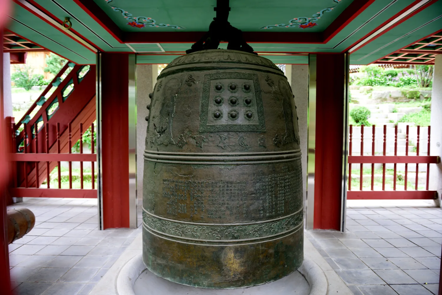 붉은 기둥과 전통 건축물 사이에 자리한 높이 2미터가 넘는 큰 청동 종의 정면 모습, 종 표면에 조각과 문자가 보인다.