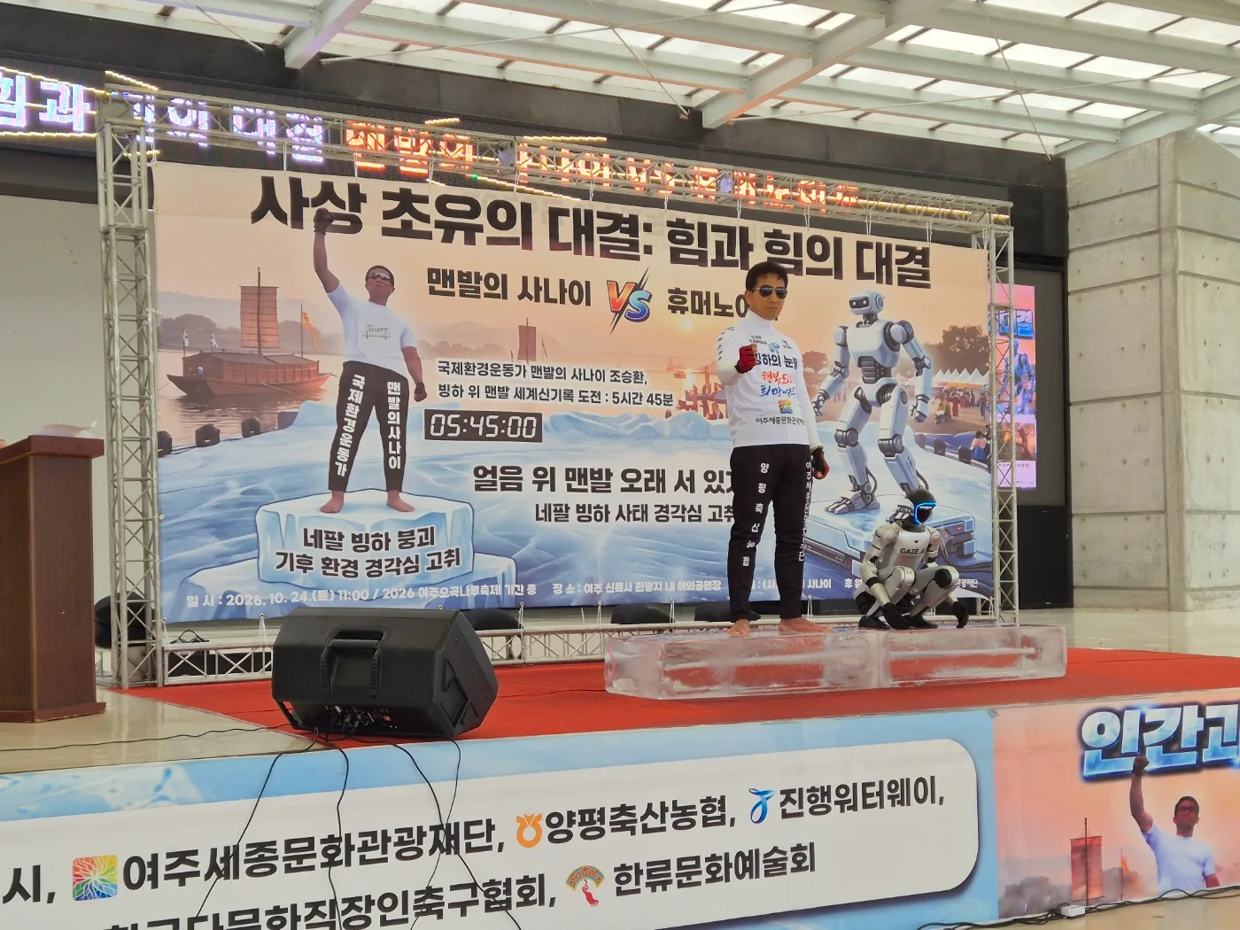 무대 위에 서서 휴머노이드 로봇과 함께 맨발로 얼음 위에 있는 조승환 씨. 무대 뒤에 대결 내용을 알리는 대형 현수막이 걸려 있음.