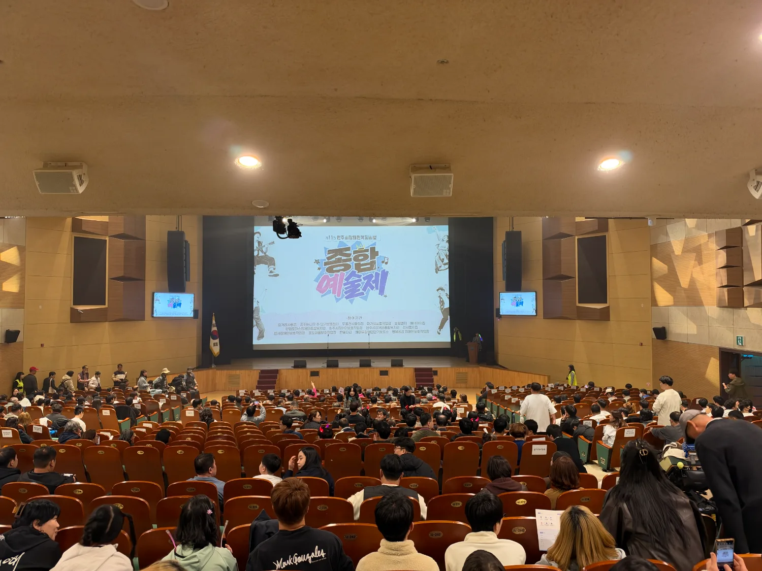 큰 공연장 객석이 가득 차 있고, 무대 스크린에 '종합 예술제' 문구가 표시되어 있으며 관객들이 앉아 있는 모습