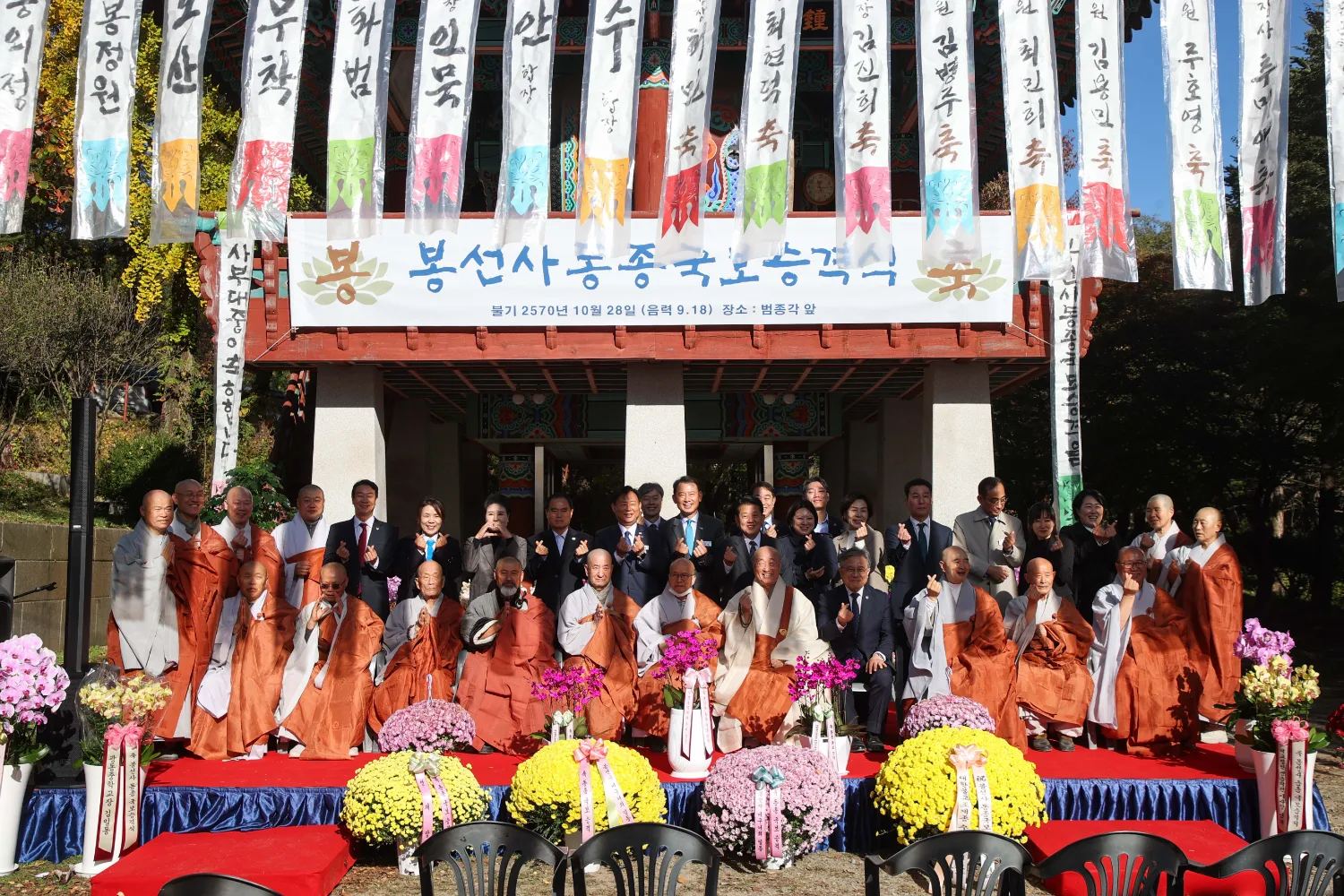 단풍이 든 나무 아래에서 주황색 승복과 정장 차림의 인물들이 무대 위에 모여 단체 사진을 찍는 모습.