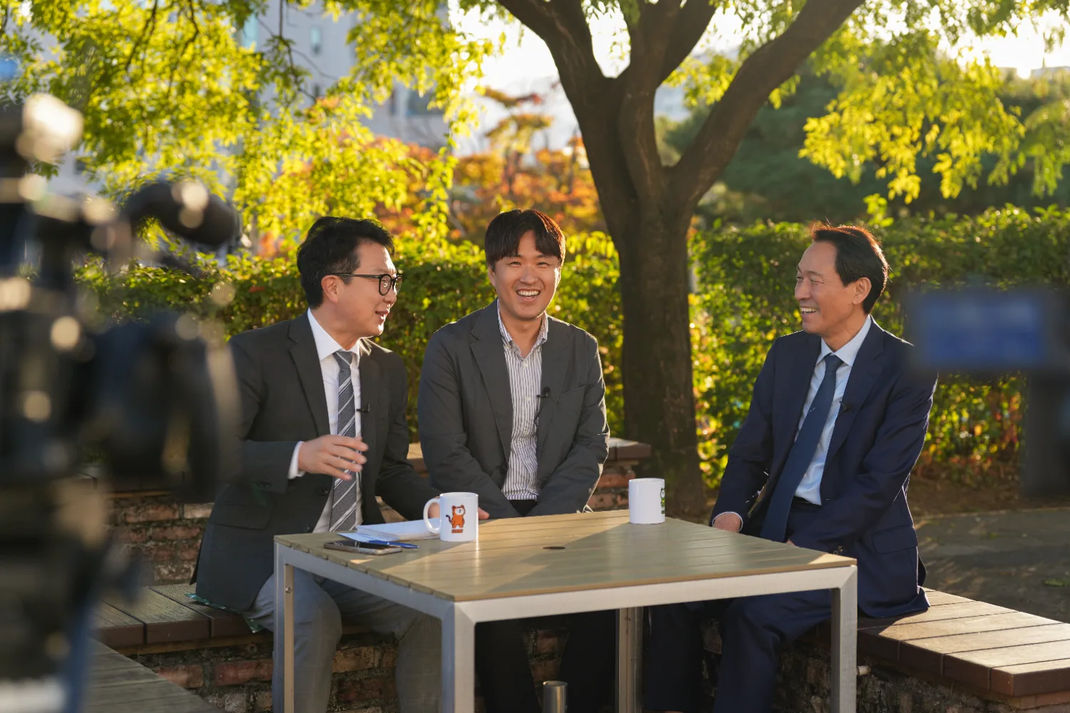 가을 분위기의 야외 벤치에 앉아 정장 차림의 세 남성이 웃으며 대화하는 모습, 앞에 테이블이 놓여 있다.