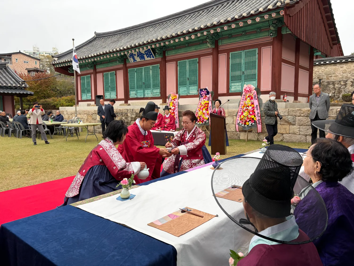 원주향교 마당에서 전통 한복을 입은 행사진행자가 어르신에게 술잔을 드리며 존경을 표하는 장면.