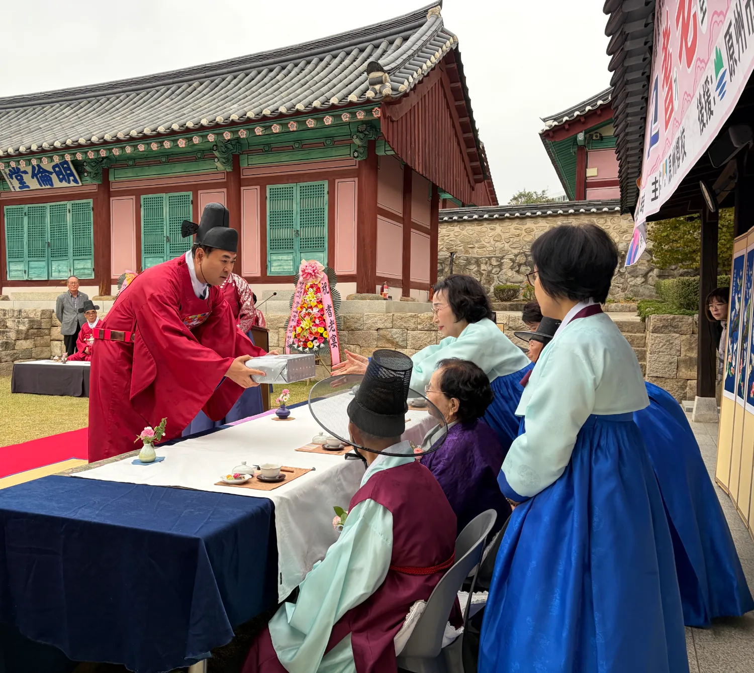 전통 복장을 한 행사진행자가 원주향교 마당에서 어르신들에게 선물 상자를 건네며 인사하는 장면.