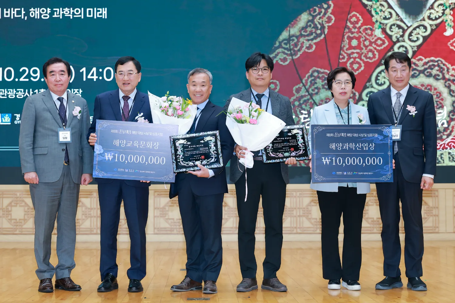 수상자와 관계자 여섯 명이 무대 위에서 나란히 서서 상패와 꽃다발을 들고 기념 사진을 찍는 모습.