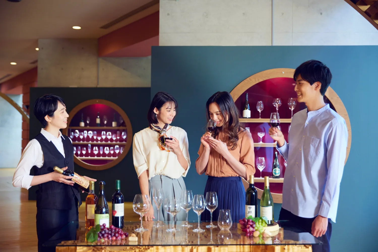 여러 사람이 와인잔을 들고 와인 향을 맡으며 즐거운 표정을 짓고 있는 실내 장면, 테이블 위에는 와인병과 포도가 놓여 있다.