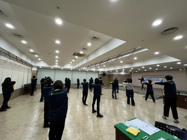 넓은 강당에서 마스크를 쓴 여러 명의 직장인이 일정 간격을 두고 서서 앞을 바라보며 자세 교정 운동을 하고 있다.